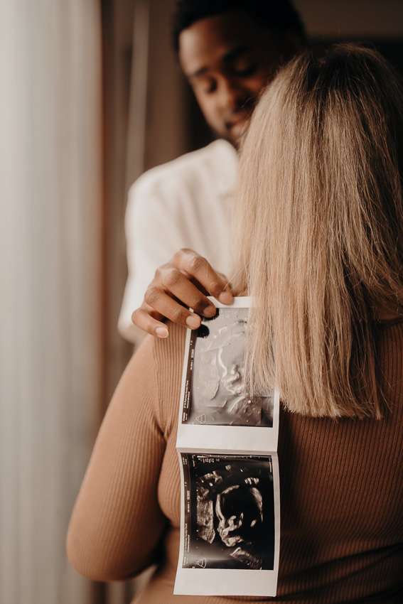 Schwangere Frau und ihr Partner umarmen sich, halten Ultraschallbild in die Kamera