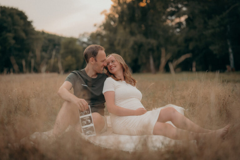 Babybauchfotos beim Schwangerschaftsshooting in Langen – Paar sitzt im Gras bei Sonnenuntergang